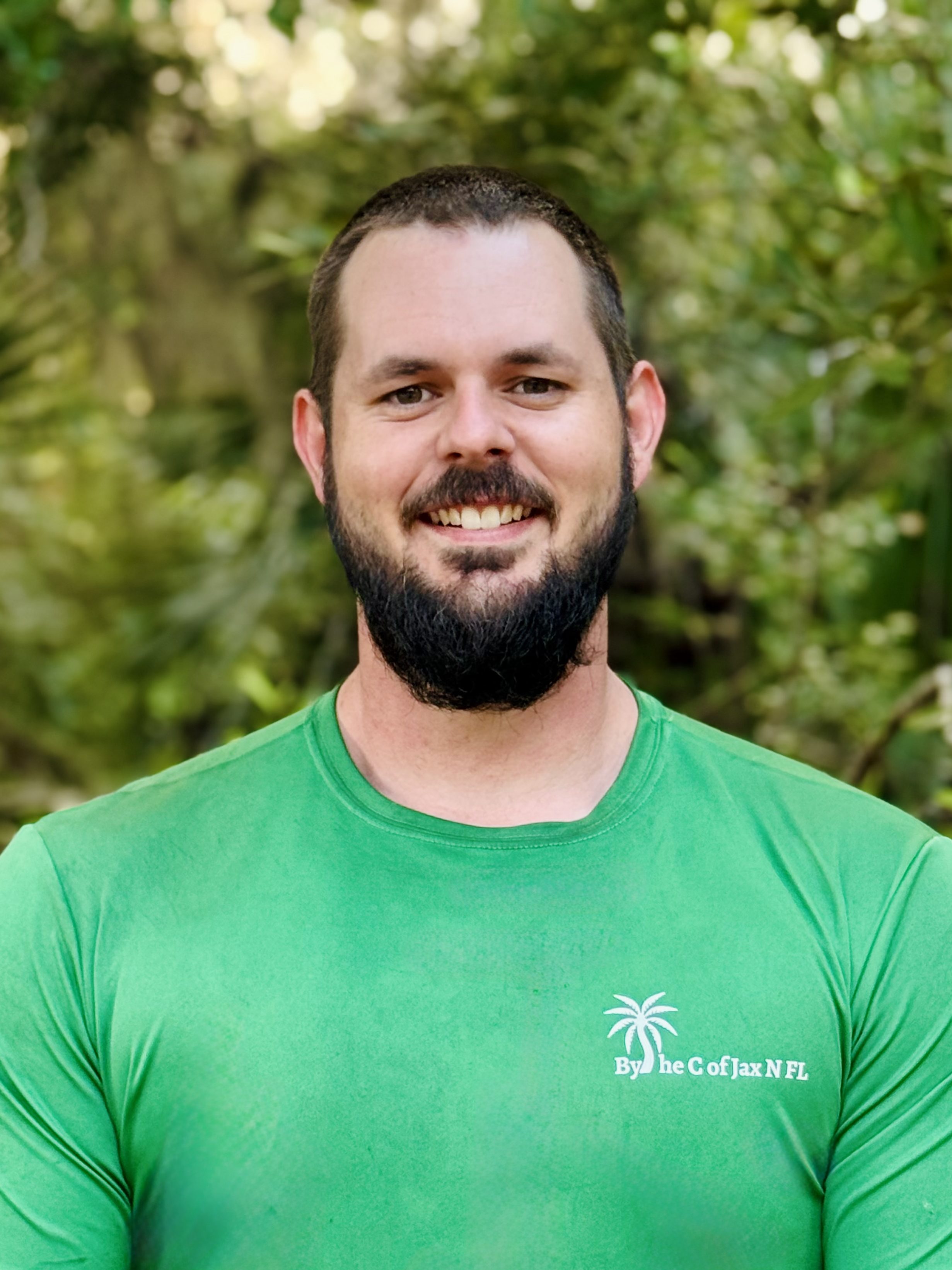 A smiling man with a beard is wearing a green shirt in a natural outdoor setting with trees and foliage in the background.