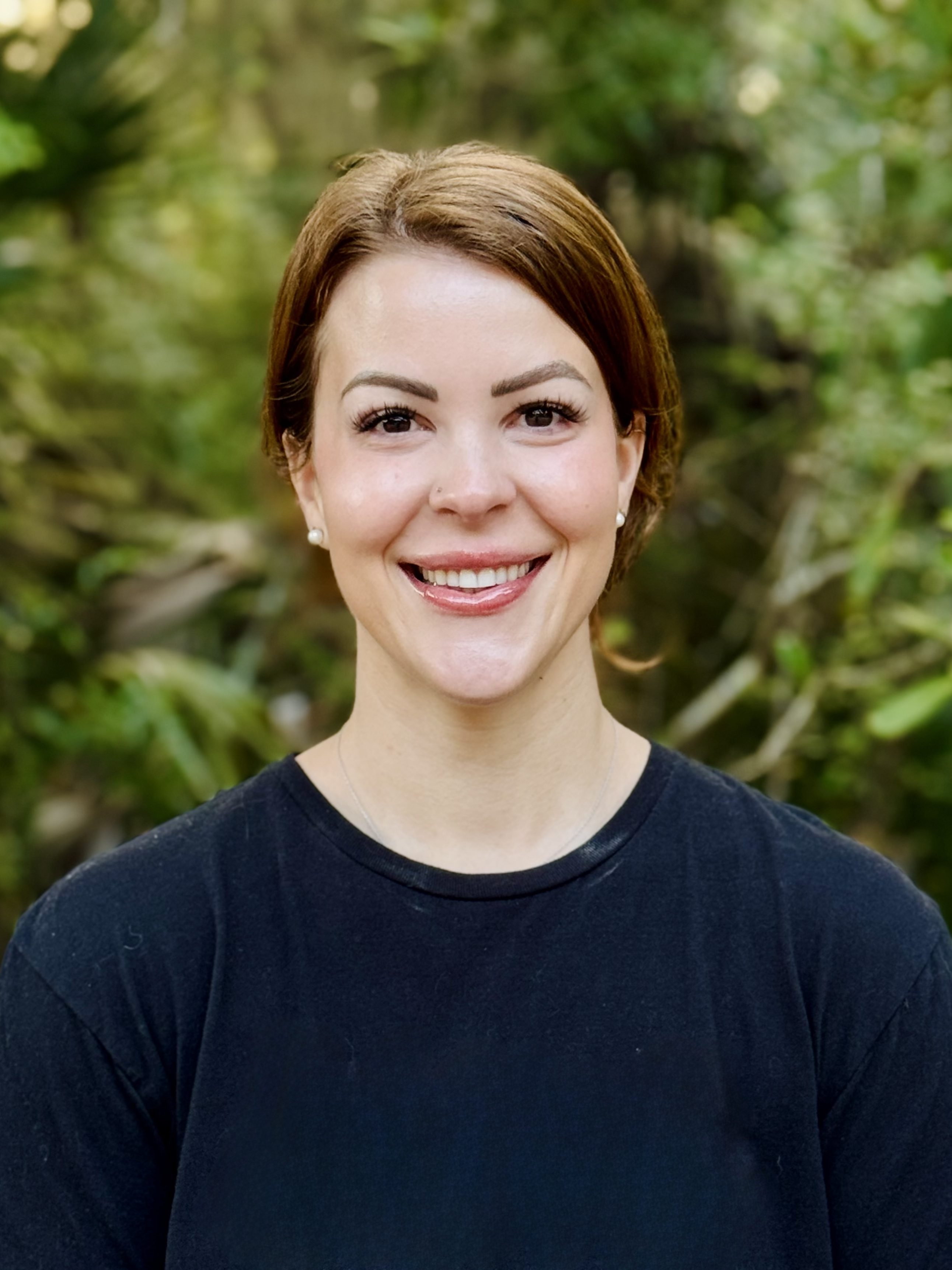 A person smiling in a black t-shirt, standing outdoors with greenery in the background.